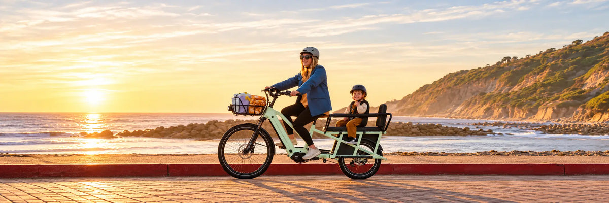 A woman rides a large cargo bicycle along a beach boardwalk at sunset, with two children seated in the rear cargo area. The scene includes the ocean, rocky shoreline, and a sunlit hillside in the background.